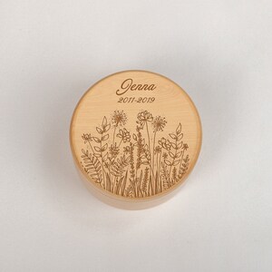 Op de afbeelding: Een ronde houten doos met een bloemenmotief en de naam "Jenna" en de data "2011-2019" gegraveerd op de deksel.