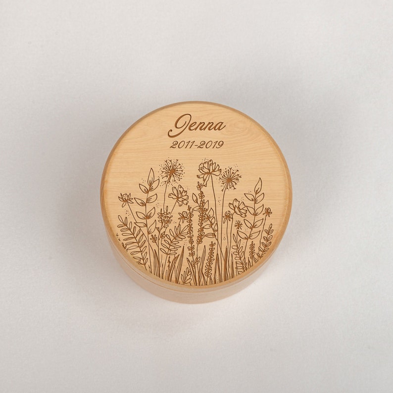 Op de afbeelding: Een ronde houten doos met een bloemenmotief en de naam "Jenna" en de data "2011-2019" gegraveerd op de deksel.