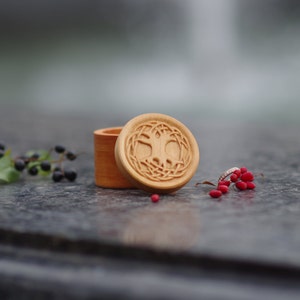 Pu&ograve; includere: Una scatola di legno per anelli con un design a forma di albero della vita intagliato sul coperchio. La scatola &egrave; aperta e un anello d'argento &egrave; visibile all'interno. La scatola &egrave; appoggiata su una superficie di pietra grigia con bacche rosse sparse intorno.