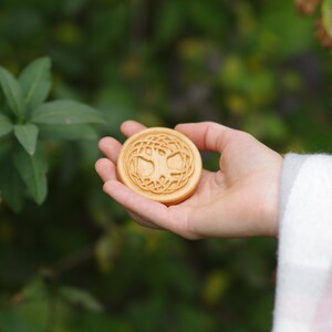 Pu&ograve; includere: Una scultura rotonda in legno di un albero della vita con intricati dettagli di nodi. La scultura &egrave; tenuta in una mano su uno sfondo verde.