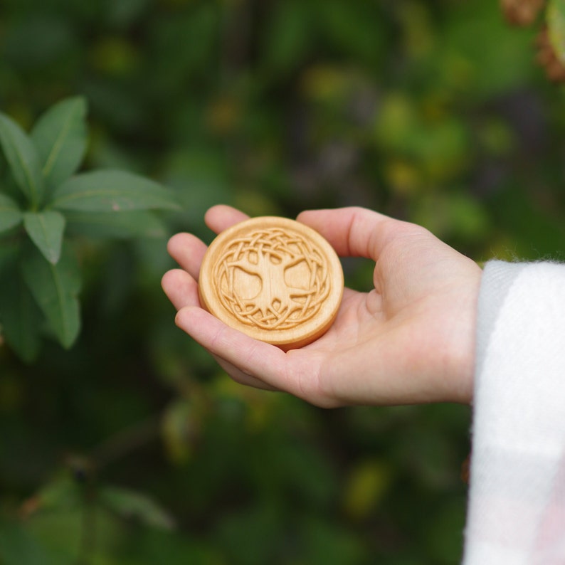 Pu&ograve; includere: Una scultura rotonda in legno di un albero della vita con intricati dettagli di nodi. La scultura &egrave; tenuta in una mano su uno sfondo verde.