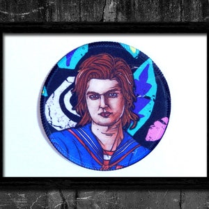 May include: A round embroidered patch featuring a character with brown hair and blue eyes wearing a blue and white sailor uniform. The background is a colorful abstract design with pink, blue, and green colors.