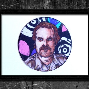 May include: A round, colorful, illustrated patch featuring a man with a mustache and a patterned background.