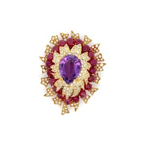 May include: A gold brooch with a large purple amethyst gemstone surrounded by smaller diamonds and red rubies.