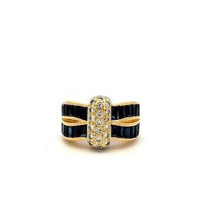 May include: A gold ring with a central vertical section covered in small, round, clear stones. The ring has two horizontal bands of dark blue rectangular stones on each side of the central section. The ring is set against a white background.