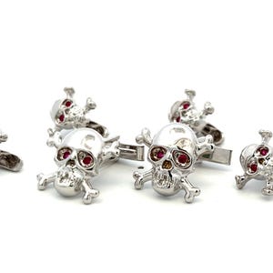 May include: A set of silver-toned metal cufflinks featuring a skull and crossbones design. Each skull has two small, circular red stones set into the eye sockets. The cufflinks are polished and reflective, showing a metallic finish throughout the structure.