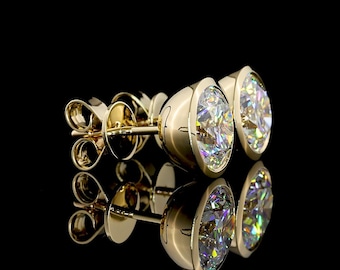 May include: A pair of stud earrings featuring round, brilliant-cut clear stones set in polished yellow gold bezels. The earrings include matching gold post backs and are displayed against a black background with a clear reflection visible below.