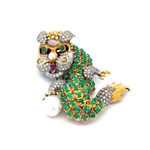 May include: A detailed brooch in the shape of a mythical creature. The brooch features a head with diamond and gold accents, emerald eyes, and a pearl. The body is adorned with emeralds and gold, with a diamond-studded tail.