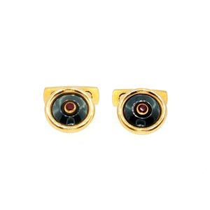 May include: A pair of gold-toned cufflinks featuring a circular design with a polished black inlay and a small, centered red gemstone. The metal frame is smooth and rounded, providing a classic look for these circular accessories.