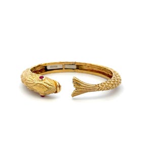 May include: A gold cuff bracelet shaped like a fish with textured scales along the band. The head of the fish features a small, round red gemstone for an eye. The bracelet has an open design, with the fish head on one end and a tail on the other.