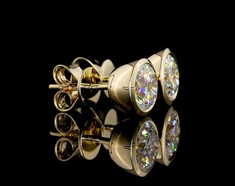 May include: A pair of gold stud earrings featuring round, brilliant-cut clear stones set in a smooth, polished gold bezel setting. The earrings include secure gold post backs and are displayed against a solid black background with a clear reflection.