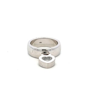 May include: A silver ring with a wide band and a heart-shaped charm. The charm is suspended from the ring and appears to have a clear stone in the center. The ring is set against a white background.