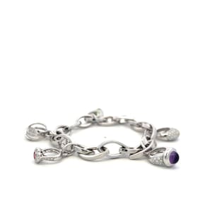 May include: A silver-toned metal charm bracelet featuring multiple dangling ring-shaped charms. The charms include a purple cabochon stone, a small red stone, and several pavé-set clear stones on polished silver-colored metal links.