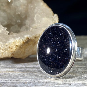 May include: A silver ring with a black oval gemstone set in a bezel setting. The gemstone has a sparkly, starry appearance.