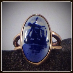 May include: A gold ring with an oval blue and white ceramic stone featuring a silhouette of a windmill and a ship.