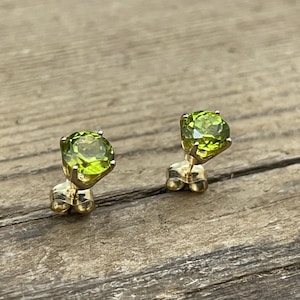 Puede incluir: Un par de pendientes de botón con piedras preciosas de peridoto redondas y facetadas engastadas en oro. Las piedras de peridoto son de un color verde claro y vibrante. Los pendientes se exhiben sobre una superficie de madera.