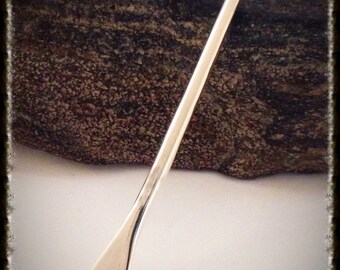 May include: A silver oar pendant, a small, detailed replica of a traditional wooden oar. The pendant is shown against a brown, textured background.