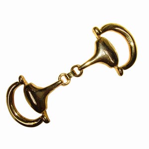 May include: A gold-colored horse bit, a piece of equestrian equipment, is displayed against a white background. The bit has a central mouthpiece with curved D-rings on each side, connected by small rings. The metal has a shiny finish.