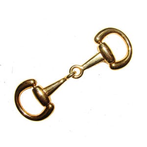 May include: A gold-toned metal horse bit buckle with a simple design. The buckle is made of two interlocking rings, each with a rounded end.