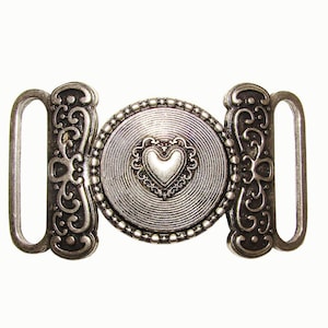 May include: Silver-toned metal belt buckle with a heart-shaped center. The buckle features a circular design with a heart in the middle, surrounded by a beaded border. The sides have decorative scrollwork and loops for attachment.