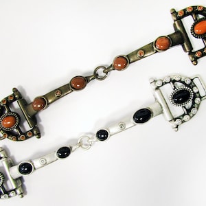 May include: Two metal belt buckles with decorative details. One buckle is gold-toned with orange stones and the other is silver-toned with black stones.