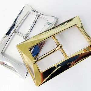 May include: Two belt buckles, one silver and one gold, with a rectangular shape and a slightly curved edge. The buckles are both polished and shiny.