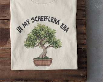 May include: Cream-colored t-shirt featuring a watercolor illustration of a bonsai tree in a brown pot. The text "IN MY SCHEFFLERA ERA" arches above the tree. The shirt is folded on a wooden surface, with white sneakers visible.