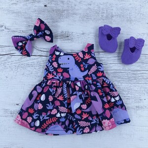 May include: A navy blue and purple dinosaur-themed dress with matching bow and shoes. The dress features a dinosaur print with pink and purple floral accents. The shoes are solid purple with bow details. The bow is navy blue with a purple dinosaur print.