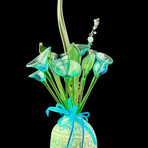 May include: A decorative arrangement of translucent green glass flowers and leaves in a round, textured light green vase. The vase features a lace-like pattern and is tied with a bright blue ribbon. A thin, spiraled wire with small beads extends upward.