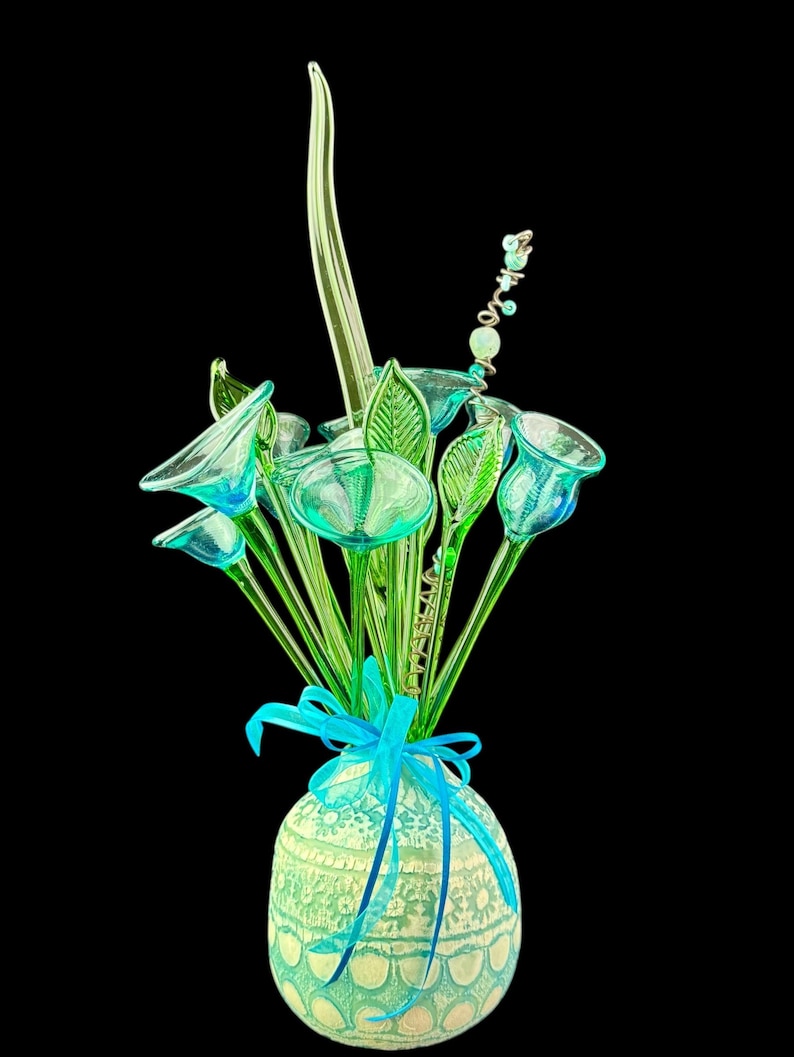 May include: A decorative arrangement of translucent green glass flowers and leaves in a round, textured light green vase. The vase features a lace-like pattern and is tied with a bright blue ribbon. A thin, spiraled wire with small beads extends upward.