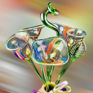 May include: A glass sculpture featuring multiple trumpet-shaped flowers with swirling patterns of blue, orange, and white. The flowers are attached to green glass stems, with one stem curving upward into a loop. A green glass leaf is attached to one side.