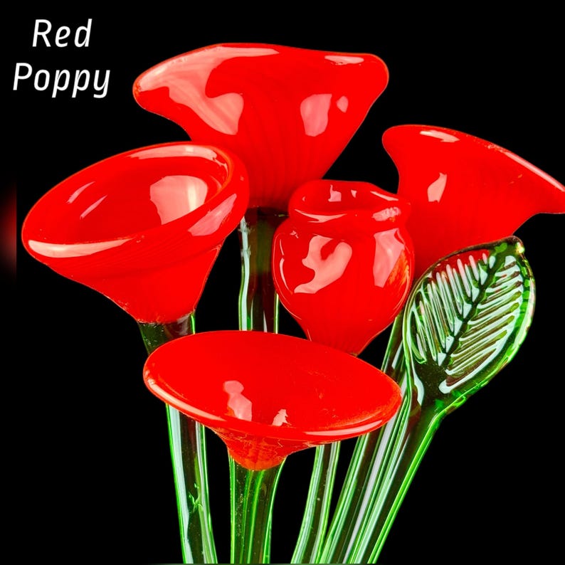 May include: A collection of glass flowers featuring vibrant red poppy blossoms with smooth, rounded petals and translucent green stems. One prominent green glass leaf with detailed veining is visible. The text Red Poppy is displayed.