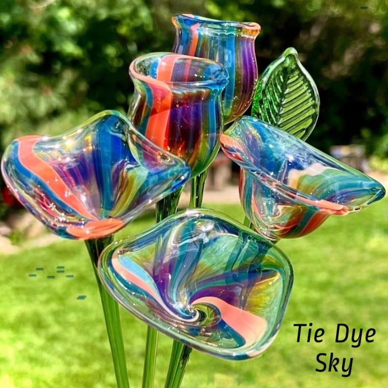May include: A collection of glass flowers featuring a tie-dye sky color pattern with swirls of blue, purple, orange, and clear glass. The set includes four individual flower-shaped glass heads on thin green stems and one green glass leaf.