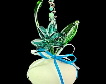 May include: A decorative egg with a teal glass flower and green leaves. The flower has a spiral accent and is tied with white and blue ribbons. The egg is off-white and set against a black background.