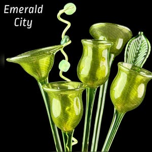 May include: A collection of green glass flower sculptures with a spiral wire accent featuring small beads. The text Emerald City is visible in the upper left corner. The glass flowers have translucent, textured petals and stems against a black background.