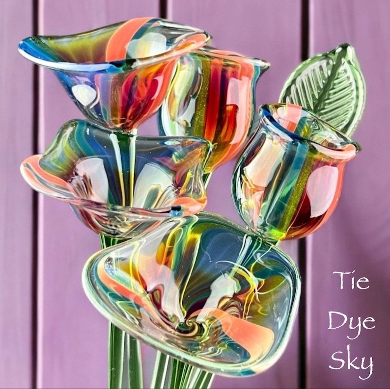 May include: A bouquet of hand-blown glass flowers featuring swirling tie-dye patterns in blue, orange, yellow, and green. The glass petals have a translucent finish with visible stems and a single green glass leaf. The text Tie Dye sky is visible.