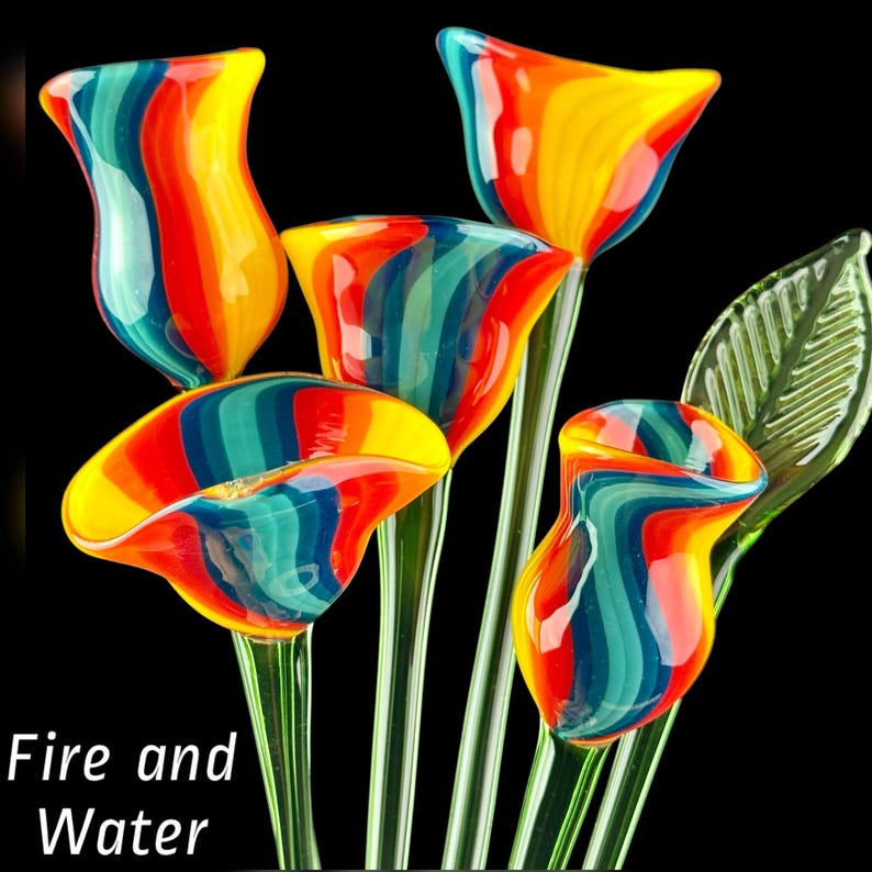 May include: A collection of five hand-blown glass flowers featuring vibrant vertical stripes of red, orange, yellow, and blue. Each flower sits atop a slender green glass stem, with one translucent green leaf attached. The text Fire and Water is visible.