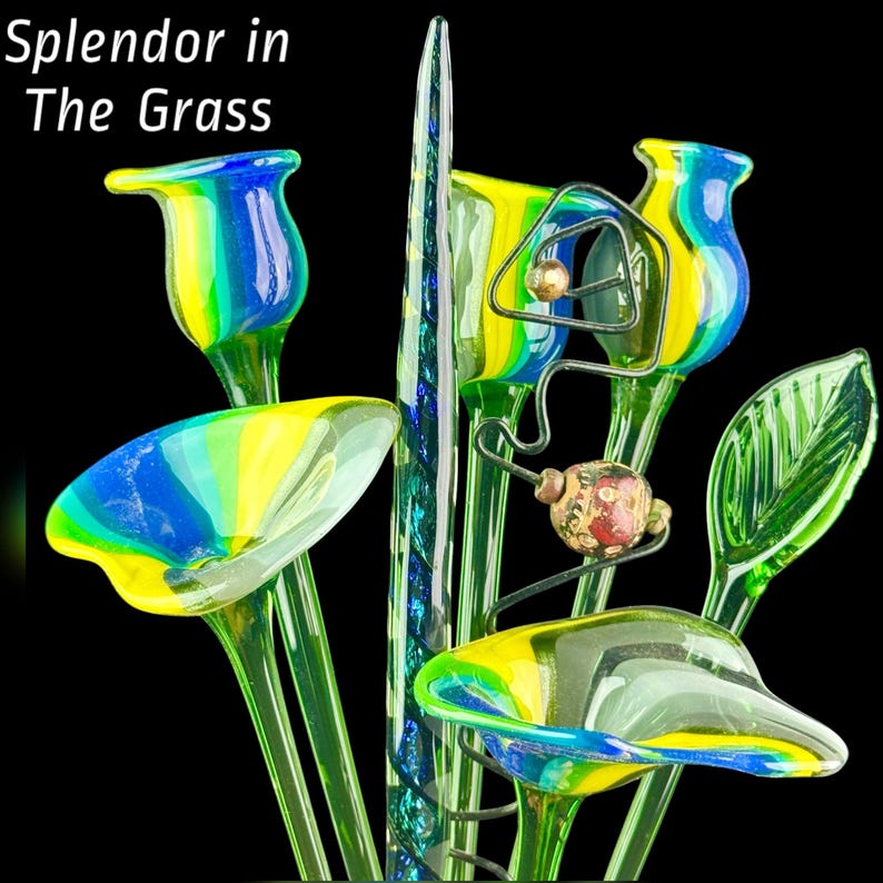 May include: A collection of glass art pieces featuring the text Splendor in The Grass. The arrangement includes blue and yellow striped trumpet-shaped flowers, a tall green spiraled spire, a green leaf, and a wire stem with a patterned bead.