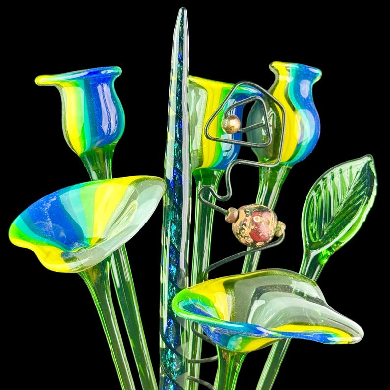 May include: A glass sculpture featuring multiple trumpet-shaped flowers with vibrant blue, yellow, and green stripes. The arrangement includes a tall, slender, spiraled glass spike, a single green glass leaf, and a dark wire accent with beads.