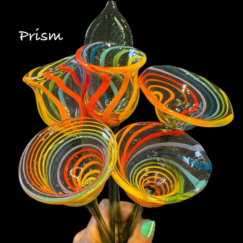 May include: A collection of five hand-blown glass flowers featuring clear glass with swirling stripes in vibrant orange, yellow, blue, and green. Each flower has a trumpet-shaped bloom attached to a green glass stem. The word Prism is visible.