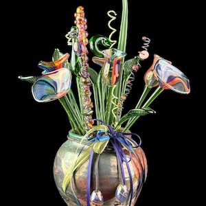 May include: A decorative arrangement of multicolored glass flowers with swirling patterns in a rounded, mottled ceramic vase. The vase is adorned with a purple and yellow ribbon tied around the neck, featuring two small glass beads hanging from the ends.