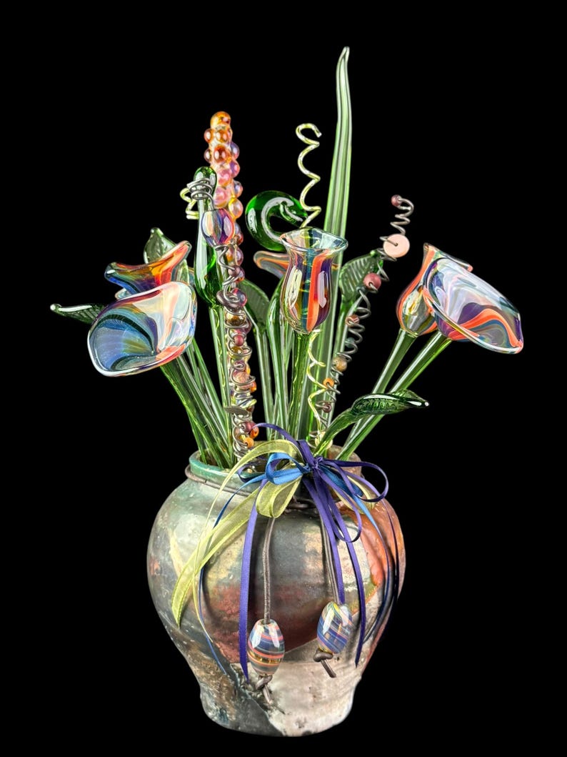 May include: A decorative arrangement of multicolored glass flowers with swirling patterns in a rounded, mottled ceramic vase. The vase is adorned with a purple and yellow ribbon tied around the neck, featuring two small glass beads hanging from the ends.