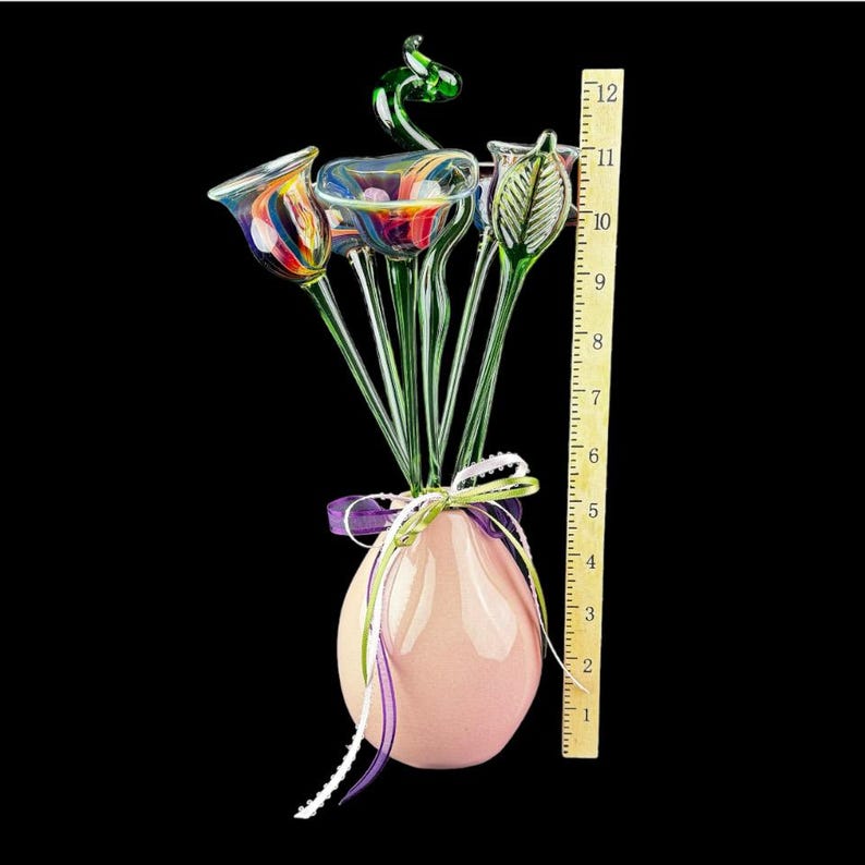 May include: A glass flower arrangement in a light pink vase, featuring multi-colored glass blossoms and green stems. A wooden ruler placed next to the vase indicates a total height of approximately 12 inches. The stems are tied with a purple ribbon.