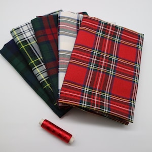 Puede incluir: Cinco piezas de tela dobladas con varios patrones de cuadros escoceses en tonos rojo, verde, blanco y azul marino, dispuestas en abanico. Un pequeño carrete de hilo de coser rojo brillante se encuentra debajo de la pila de telas.