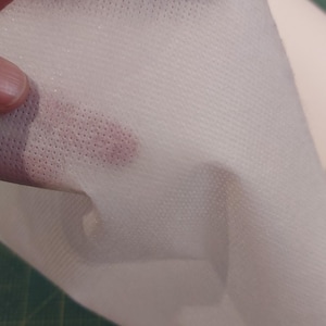 Könnte beinhalten: Nahaufnahme eines weißen, halbtransparenten Stoffes mit einem strukturierten, gepunkteten Muster. Der Stoff wird hochgehalten, wodurch seine Dünne und die Textur des Materials sichtbar werden. Der Hintergrund ist eine grüne Schneidematte.