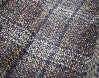 May include: Close-up of a textured fabric with a plaid pattern. The fabric features a mix of brown, tan, and white threads, with subtle red accents. The weave creates a detailed, slightly rough surface, suggesting a material suitable for outerwear.
