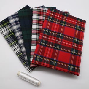Peut inclure: Un ensemble de cinq échantillons de tissu pliés présentant divers motifs à carreaux et tartan en rouge, vert, blanc, bleu marine et jaune, accompagné d'une petite bobine de fil à coudre blanc placée au bas de la pile de tissus.