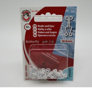 Op de afbeelding: Een verpakking met 12 witte plastic haken en ogen voor het naaien, met een afmeting van 10 mm bij 8 mm. De verpakking is rood en wit met de merknaam Koh-i-Noor en de tekst "Butterfly" erop gedrukt.