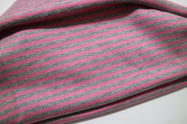 May include: A piece of fabric featuring a repeating pattern of horizontal stripes in alternating shades of heather gray and bright pink. The material has a visible knit texture with a soft, folded drape that creates subtle shadows across the surface.