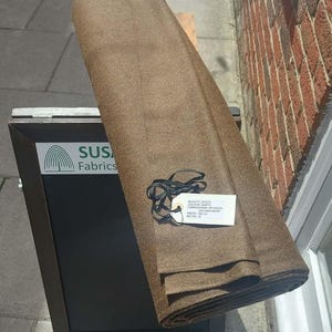 Peut inclure: Un rouleau de tissu marron avec une étiquette blanche attachée par un cordon noir. L'étiquette indique : QUALITY: GH035, COLOUR: EARTH, COMPOSITION: 80% WOOL 20% CASHMERE, WIDTH: 150 CM, METRE: 12. Le tissu repose sur un panneau noir SUS Fabrics.